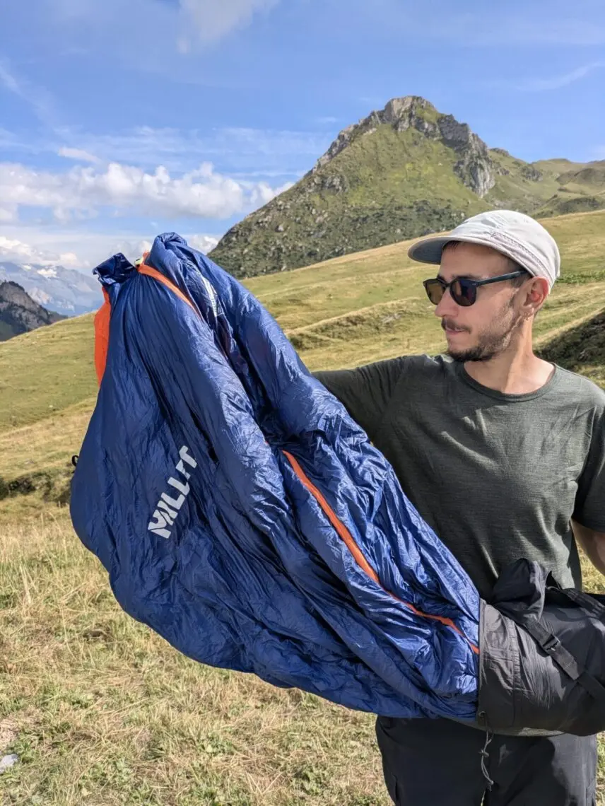 Sac de couchage en duvet déployé, tenu à bout de bras devant un paysage de montagne